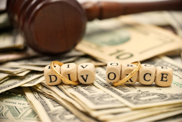 Wooden gavel, wedding rings, and letter blocks spelling 'divorce' on a pile of dollar bills.