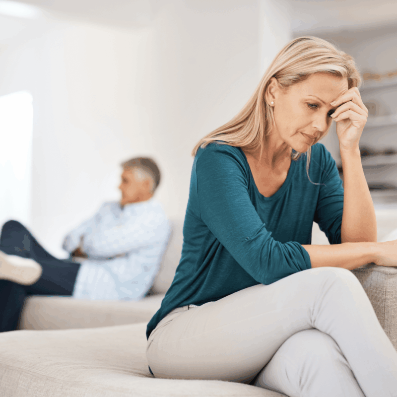 Leaving a toxic relationship. Woman at the edge of a sofa with her head in her hands with her spouse behind her on the other end of the sofa. It appears that he is not willing to listen.